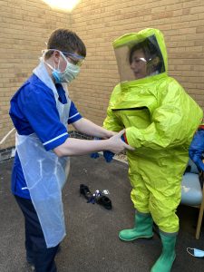 A person in a blue NHS uniform helping someone in a yellow hazard suit