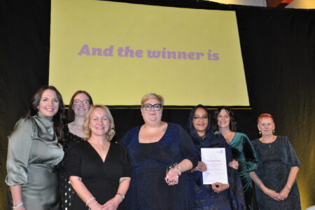 A group of smiling women standing on a stage with a large screen behind them. 