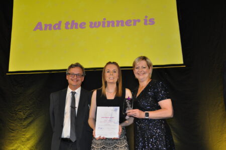 Three people smiling with a large screen behind them.