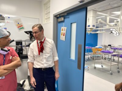 Two men stand looking into an operating theatre, one dressed in surgical scrubs.