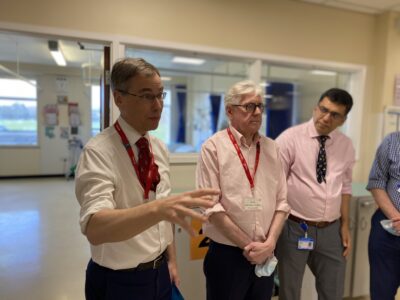 Three men stand talking, with a bay of hospital beds behind them.