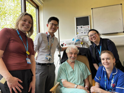 A group of people standing around a person sat in a chair, who is wearing a hospital gown.