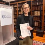 Woman from cancer team smiling holding certificate in front of a Royal College of Physicians sign