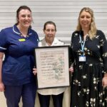 Three women standing in a line, with two in NHS uniforms. The woman in the middle is holding up a framed picture.