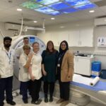 A group of people in white clinical uniforms stand with a woman in front of a CT scanner. There is a picture panel on the ceiling aove showing blue sky and palm trees.