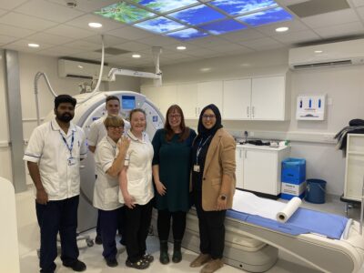 A group of people in white clinical uniforms stand with a woman in front of a CT scanner. There is a picture panel on the ceiling aove showing blue sky and palm trees.