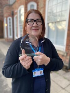 A woman with red hair and black glasses, wearing a nursing uniform, holds up her glass award trophy.