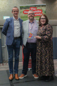 Three people, two men and a woman, stand in a line on a stage. The woman is holding an award.