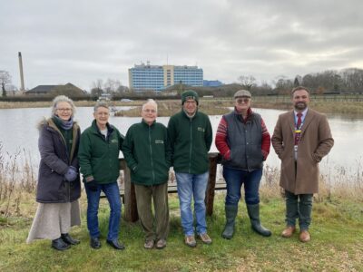 Boston Woods Trust Trustee Claire Elwood, Secretary Frances Brooks, President Adrian Isaac, Chairman Steve Elwood and Volunteer Simon Horniblow with United Lincolnshire Hospitals Charity Manager Ben Petts. They are stood on the edge of the lake with Pilgrim hospital in the background.