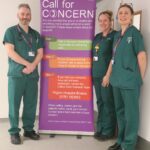 A man wearing green scrubs stands to the left of a banner about the Call for Concern Service. Two women, also wearing green scrubs, stand side-by-side to the right of the banner.