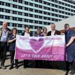 Seven people stand in a line holding a large pink and white flag. The flag has a pink heart in the centre alongside the words organ donation let's talk about it.