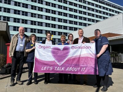 Seven people stand in a line holding a large pink and white flag. The flag has a pink heart in the centre alongside the words organ donation let's talk about it.