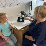 A young girl wiht blonde hair sits in a chair next to a female nurse.