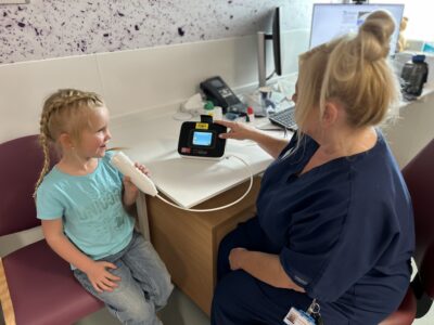 A young girl wiht blonde hair sits in a chair next to a female nurse. 