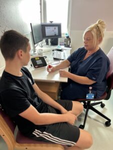 A teenage boy wearing a black t-shirt sits in a chair to the left facing a female nurse. The nurse is holding a red and white item in her hand .