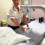A woman, who has her brown hair tied up and is wearing glasses, stands facing the camera with the DEXA scanner in front of her. She is positioning a small box on the couch where patients would lay, and a white arm from the scanner is above the box. A red laser is coming down from the scanner onto the box.