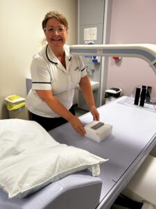 A woman, who has her brown hair tied up and is wearing glasses, stands facing the camera with the DEXA scanner in front of her. She is positioning a small box on the couch where patients would lay, and a white arm from the scanner is above the box. A red laser is coming down from the scanner onto the box.