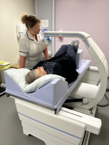 A radiographer wearing a white tunic is helping to position a woman on the couch of the DEXA scanner. 