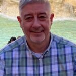 A man smiling, who is wearing a green checked shirt. He has grey hair and is facing the camera.