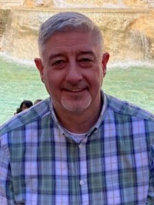 A man smiling, who is wearing a green checked shirt. He has grey hair and is facing the camera. 