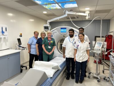 A group of six people stand in front of a large round CT scanner. 
