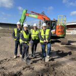 Four people stand in yellow hi-vis jackets in front of a digger inside a muddy stadium. Two people are holding spades and look as if they are preparing to dig.
