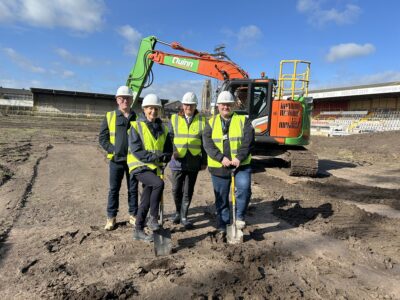 Four people stand in yellow hi-vis jackets in front of a digger inside a muddy stadium. Two people are holding spades and look as if they are preparing to dig. 