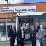 Four people are stood at the door of a bus, which has been parked outside of a building. The building has a large white sign saying Lincoln Community Diagnostic Centre. They are smiling at the camera, along with a man sat in the bus driving seat.