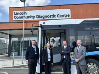 Four people are stood at the door of a bus, which has been parked outside of a building. The building has a large white sign saying Lincoln Community Diagnostic Centre. They are smiling at the camera, along with a man sat in the bus driving seat.