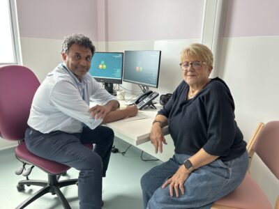 Two people sit at a table in a consultation room. There is a computer in the background. 