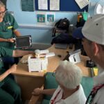 clinician in green gown demonstrating an ultrasound scanner machine to two people dressed in white