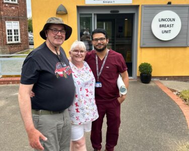A man in a green t-shirt with a lady and a doctor stood outside a yellow building