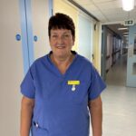 A woman wearing blue scrubs stands in a hospital corridor. She has short dark hair and is smiling.