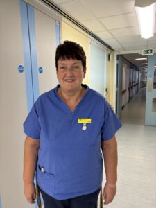 A woman wearing blue scrubs stands in a hospital corridor. She has short dark hair and is smiling. 