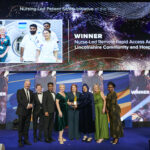 A group of people in smart clothes stand in a line underneath a large TV screen on a stage, receiving a trophy.