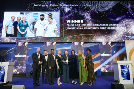 A group of people in smart clothes stand in a line underneath a large TV screen on a stage, receiving a trophy. 